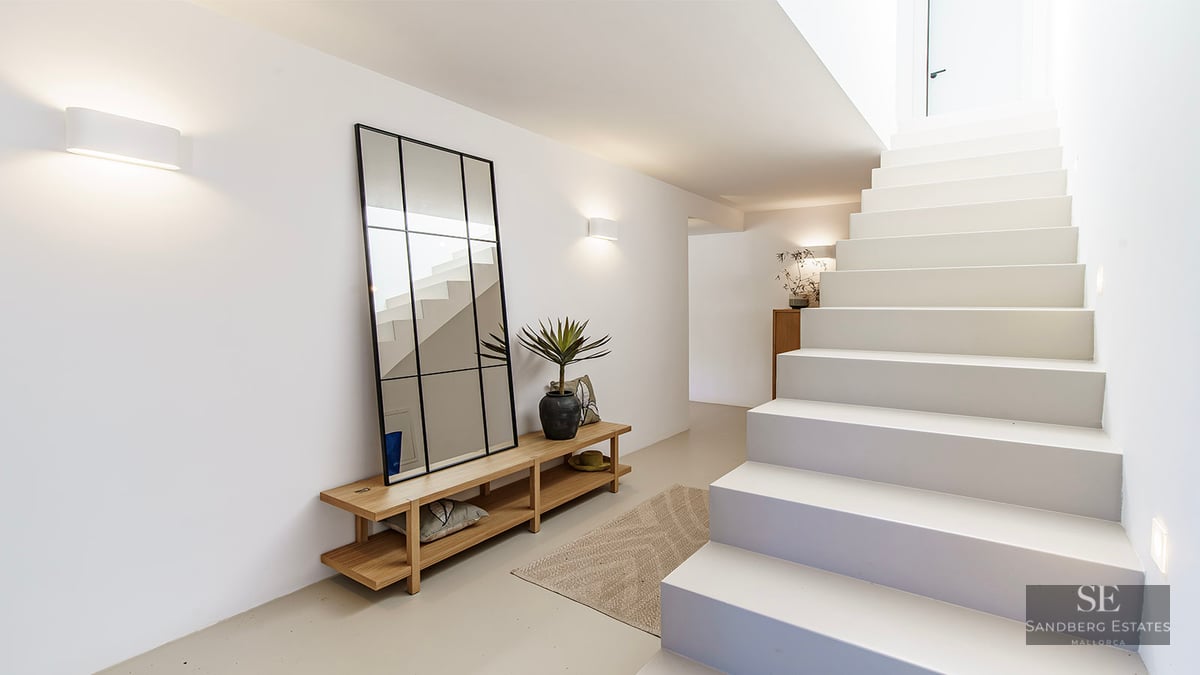 Bright minimalist hallway with white stairs, a large black-framed mirror, wooden bench, and warm wall lighting.