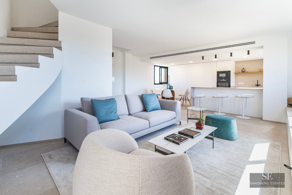 Modern living room with grey sofa, teal pillows, marble coffee table, and spiral staircase next to a white kitchen.