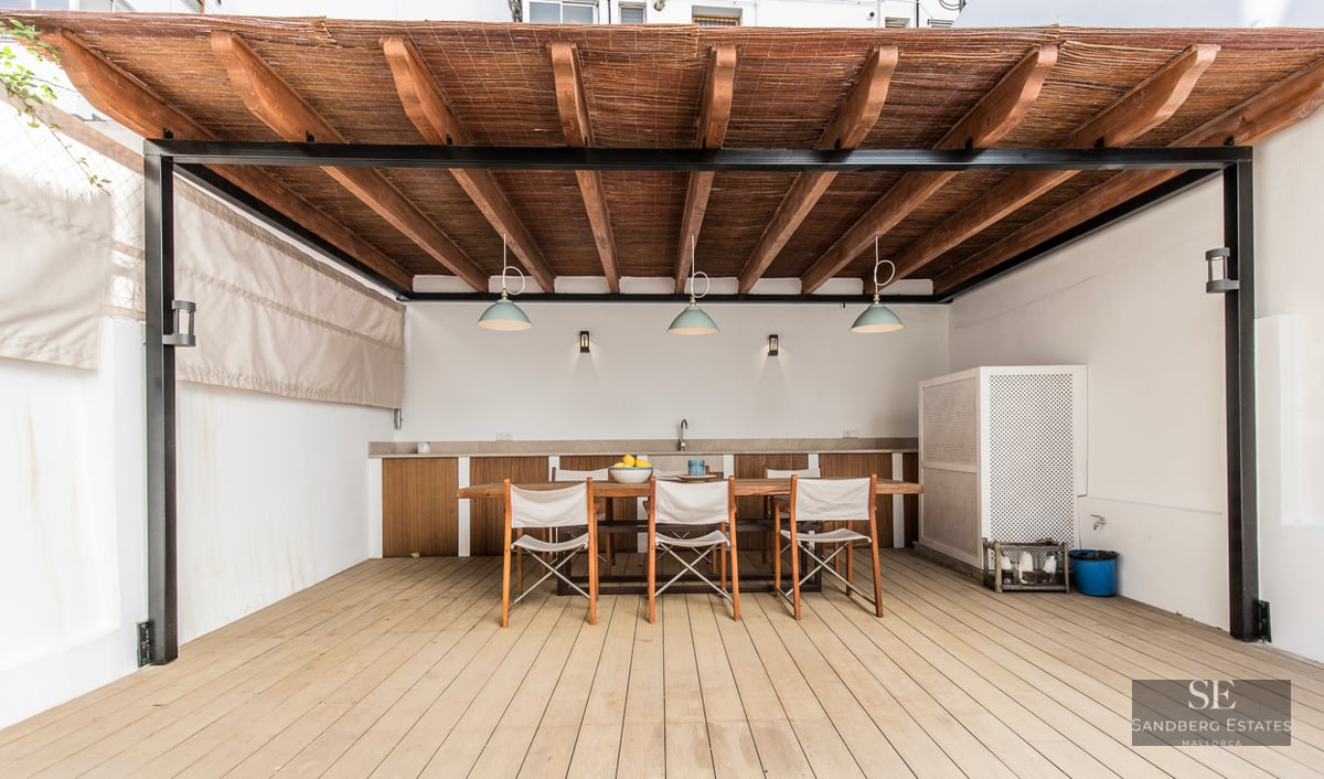 Shaded outdoor dining area with a large wooden table, chairs, and a pergola with integrated lighting and kitchenette.