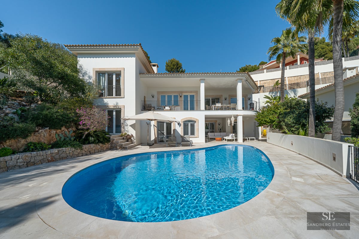 Grande piscine ovale bleu azur devant une villa blanche à deux étages avec terrasse en pierre et palmiers.