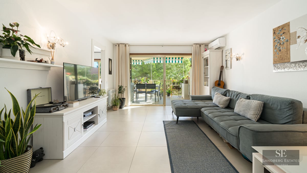 Salon moderne avec canapé bleu, meuble TV blanc et baies vitrées ouvrant sur une terrasse verdoyante.