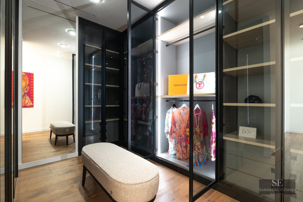 Modern walk-in closet featuring glass-door wardrobes with LED lighting, wood floors, and a textured boucle bench.