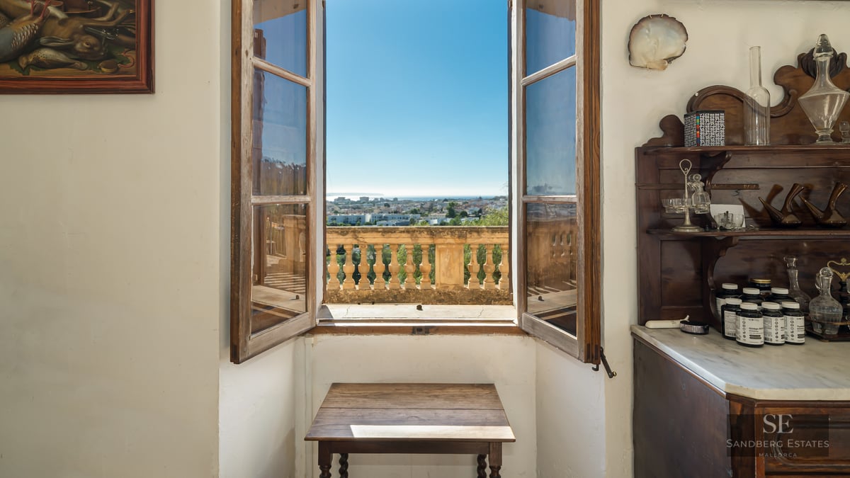 An open wooden window framing a panoramic view of a coastal city and the sea under a clear blue sky.