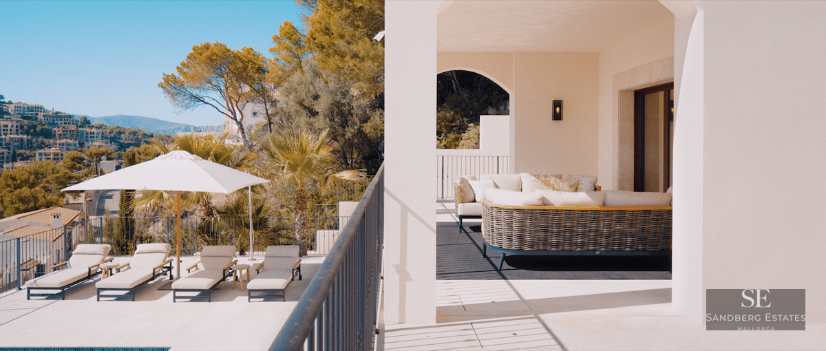 Sun loungers and a white umbrella on a stone terrace overlooking a Mediterranean hillside town.