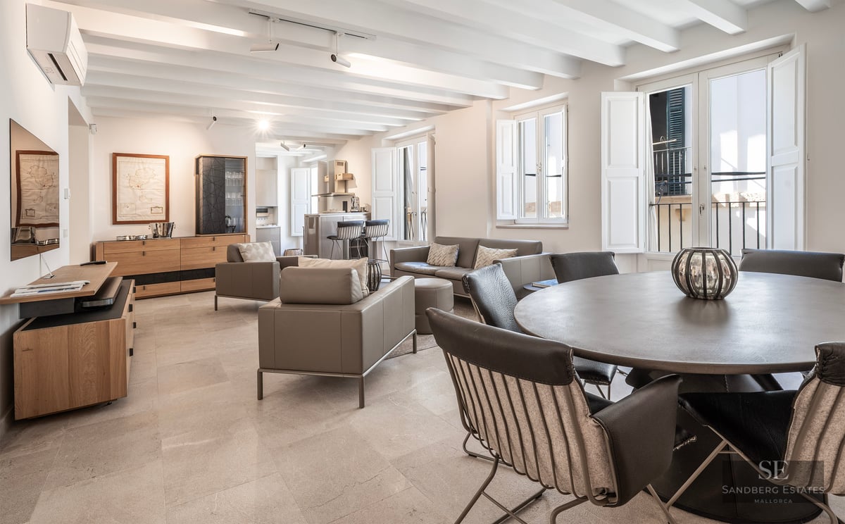 Salon spacieux avec plafonds à poutres blanches, meubles en cuir gris et table à manger ronde.