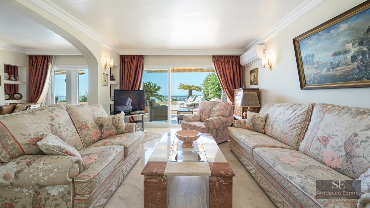 Spacious living room with floral sofas, marble table, and large glass doors opening to a terrace overlooking the sea.
