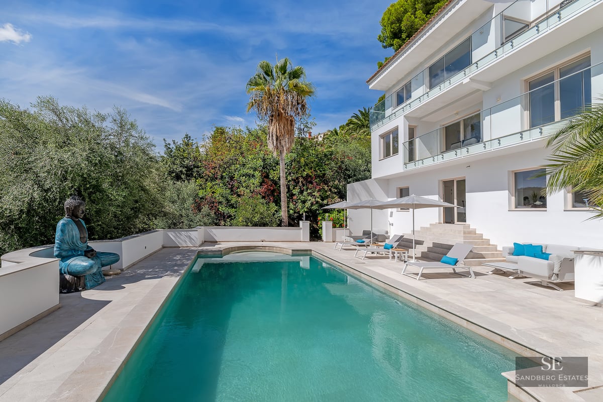 A large turquoise swimming pool next to a modern white villa with lounge chairs, umbrellas, and a Buddha statue.