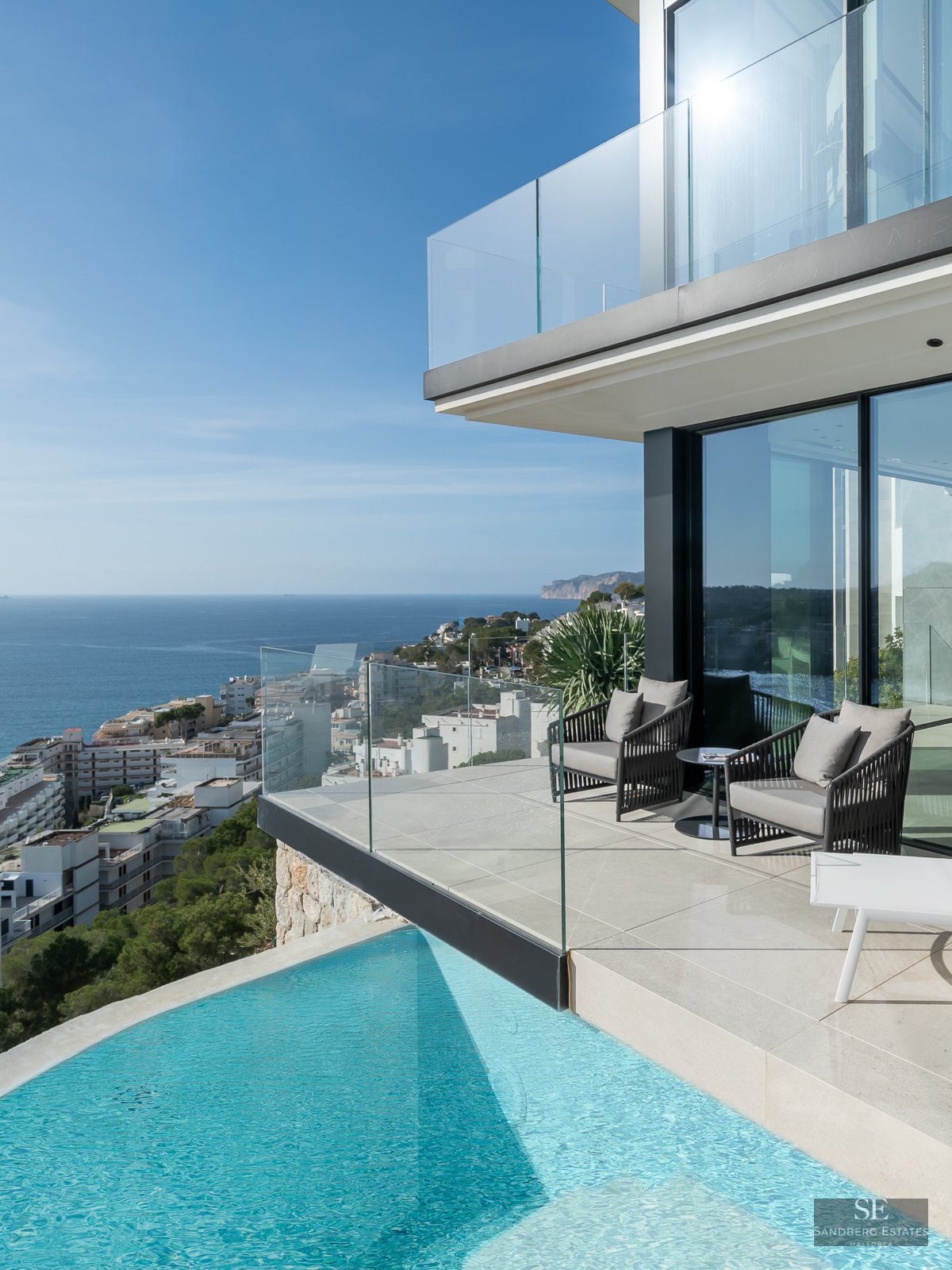 Modern infinity pool and terrace with grey armchairs overlooking the Mediterranean sea and a coastal town.