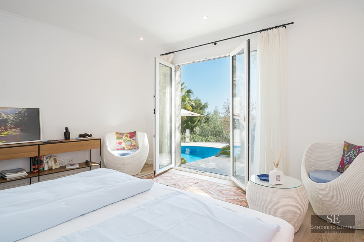Chambre lumineuse avec lit blanc et chaises design, s'ouvrant sur une piscine turquoise et un jardin.