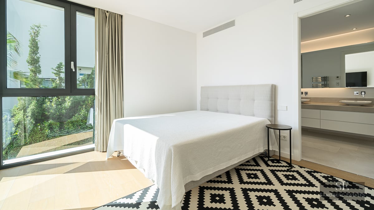 Modern bedroom featuring white bedding, a geometric rug, floor-to-ceiling window, and an ensuite bathroom.