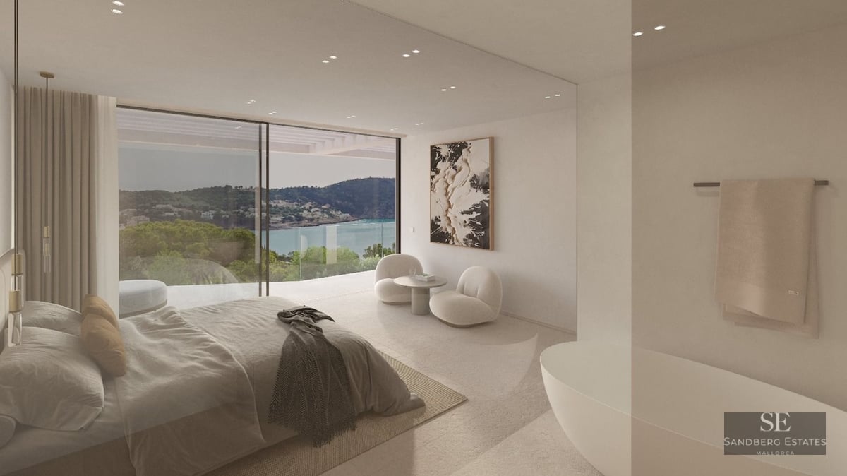Modern bedroom with floor-to-ceiling windows overlooking a bay, featuring a freestanding tub and designer furniture.