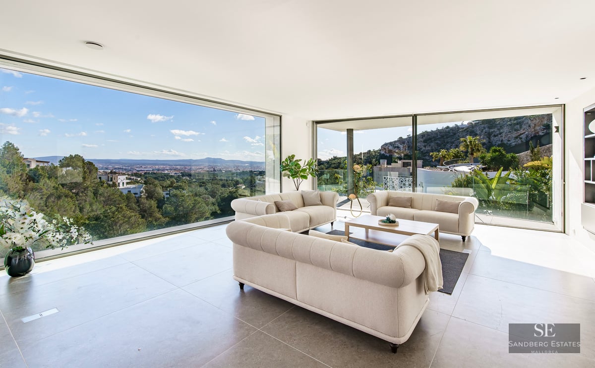 Modern living room with three beige tufted sofas and floor-to-ceiling windows overlooking a city valley and mountains.