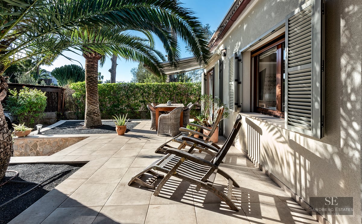 Sunny tiled patio with wooden lounge chairs, a dining set, and a large palm tree against a beige house wall with shutters.