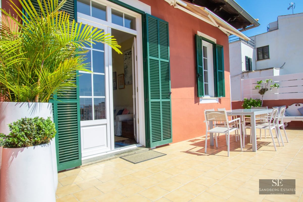 A sunny rooftop terrace featuring terracotta walls, green shutters, white dining furniture, and lush potted plants.