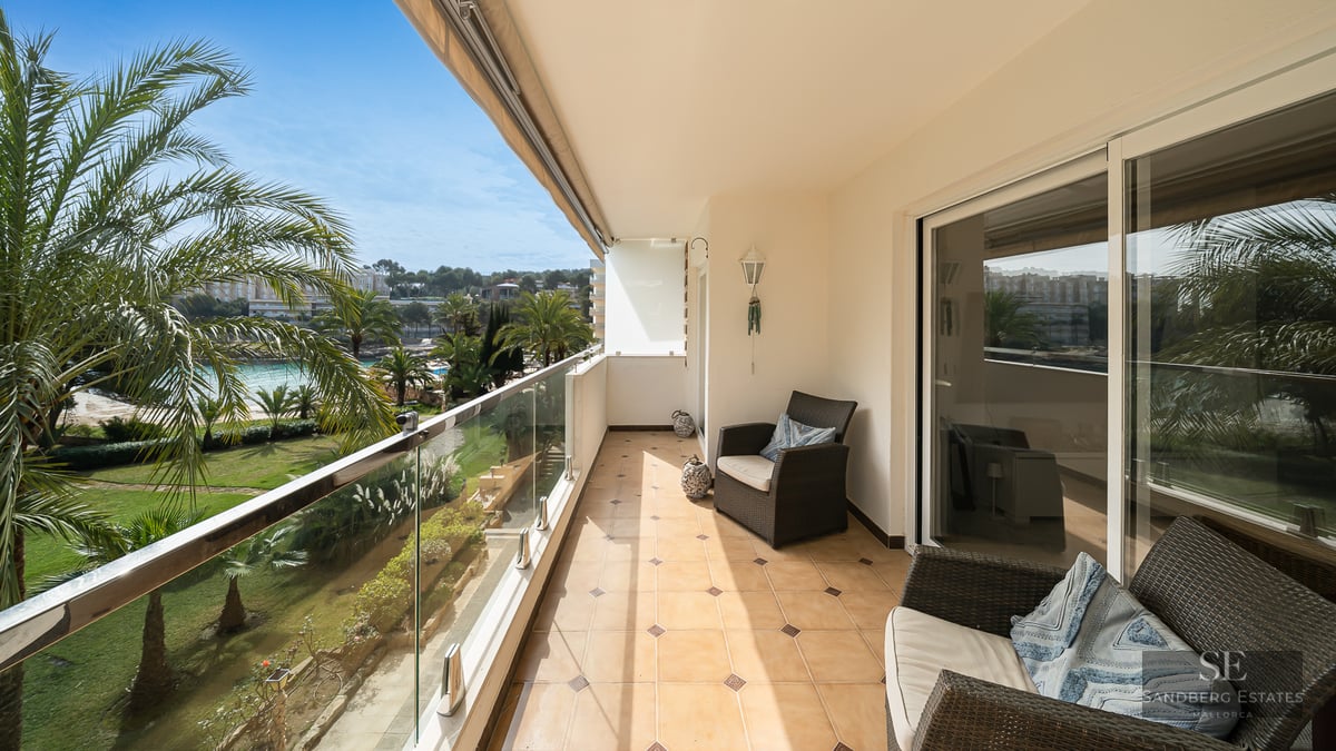 Sunlit balcony with wicker chairs, glass railing, and a scenic view of lush palm trees and a garden.