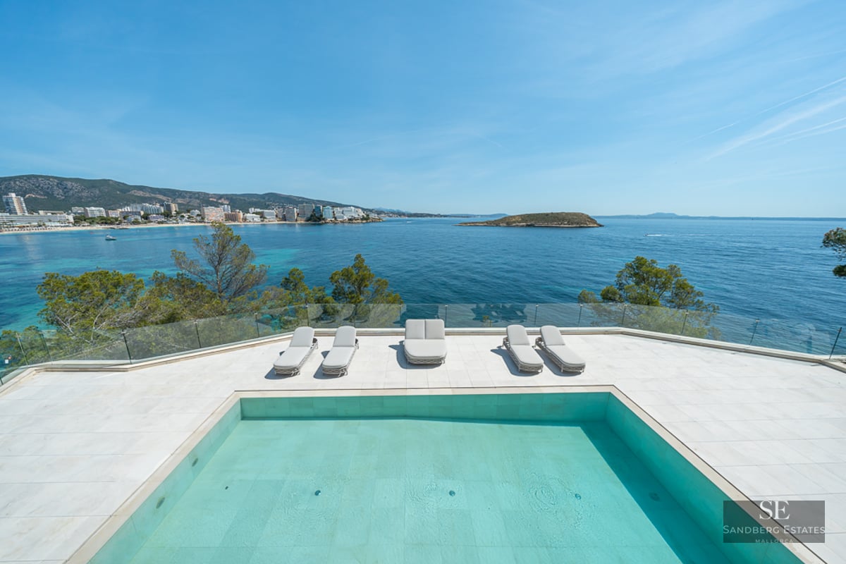 Modern swimming pool and sun loungers on a white terrace overlooking a turquoise bay and distant coastline.