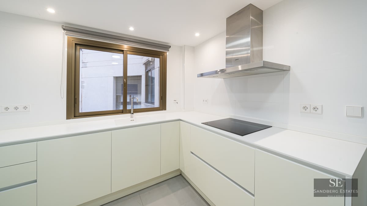 Minimalist white kitchen featuring L-shaped counter, induction stove, stainless steel extractor, and window.