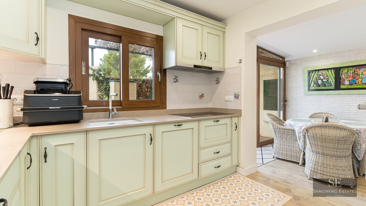 Bright kitchen with mint green cabinets, wooden window, and patterned floor leading to a dining area with wicker chairs.