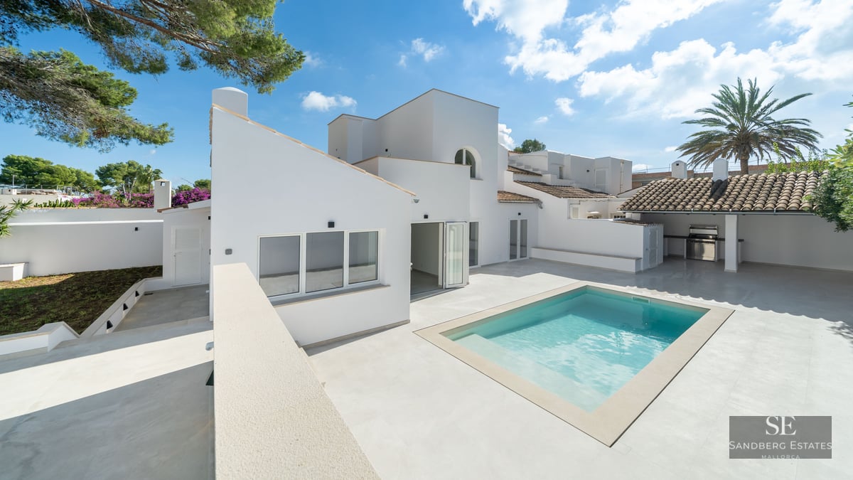 Modern white Mediterranean villa featuring a bright swimming pool, spacious stone patio, and outdoor kitchen area.