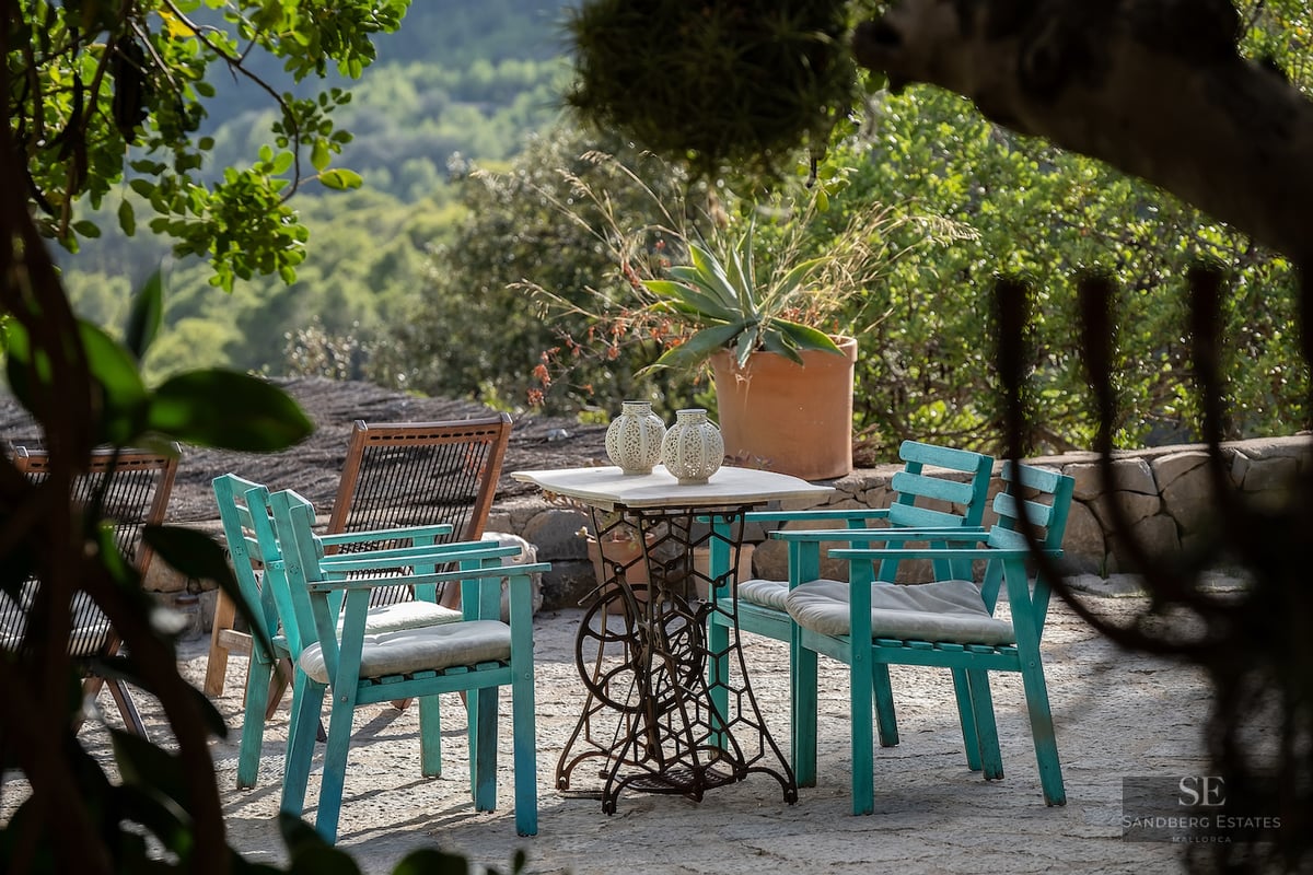 Vintage marble table and teal wooden chairs on a stone patio surrounded by lush Mediterranean greenery and mountain views.