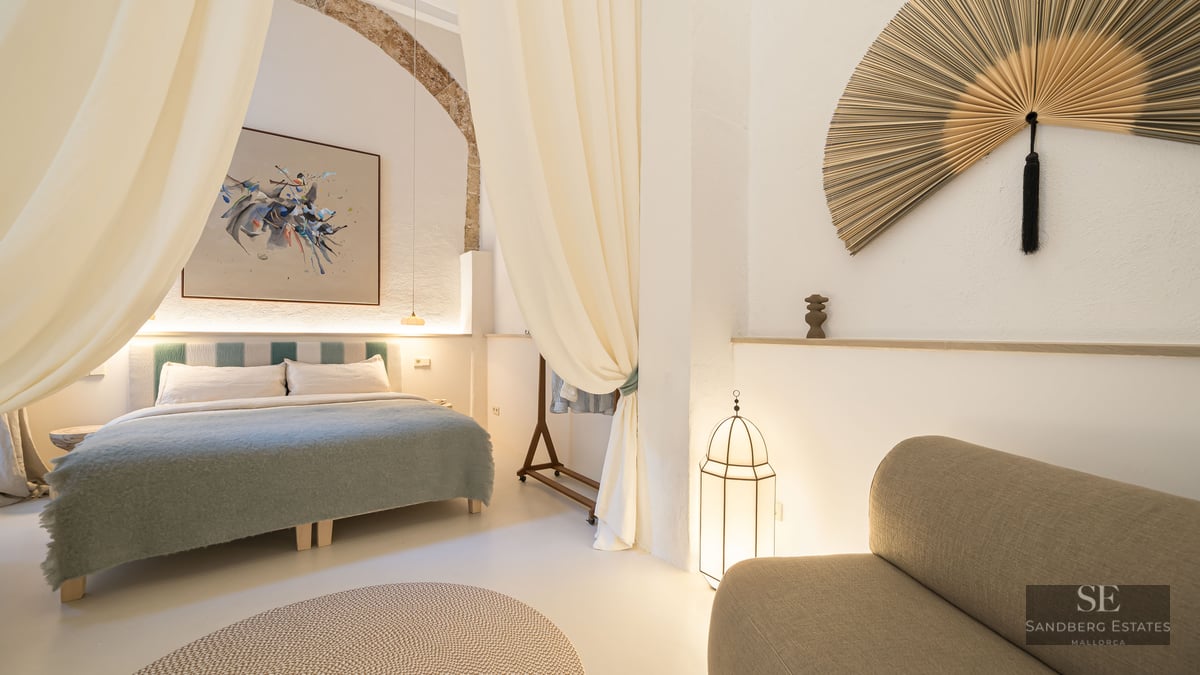 Chambre de luxe avec grand lit, arche en pierre d'origine, rideaux fluides et décor méditerranéen minimaliste.