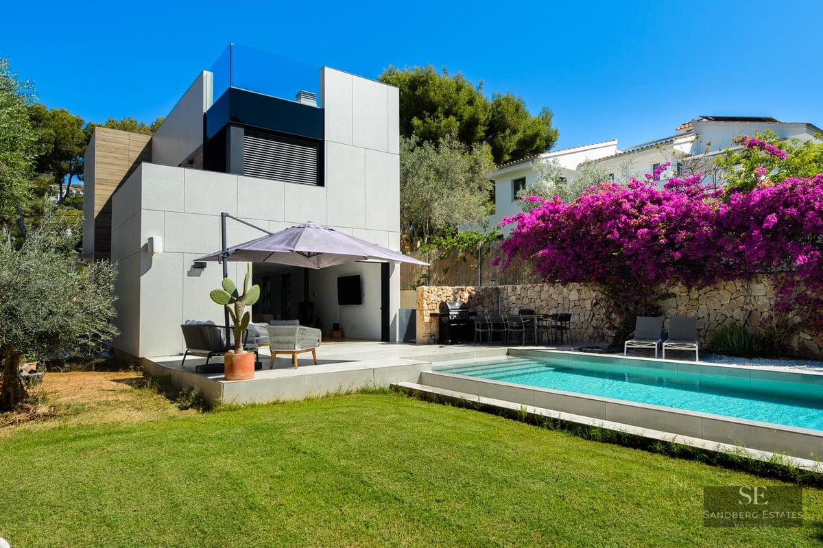 Modern villa exterior with a blue rectangular pool, green lawn, and bright purple bougainvillea against a stone wall.