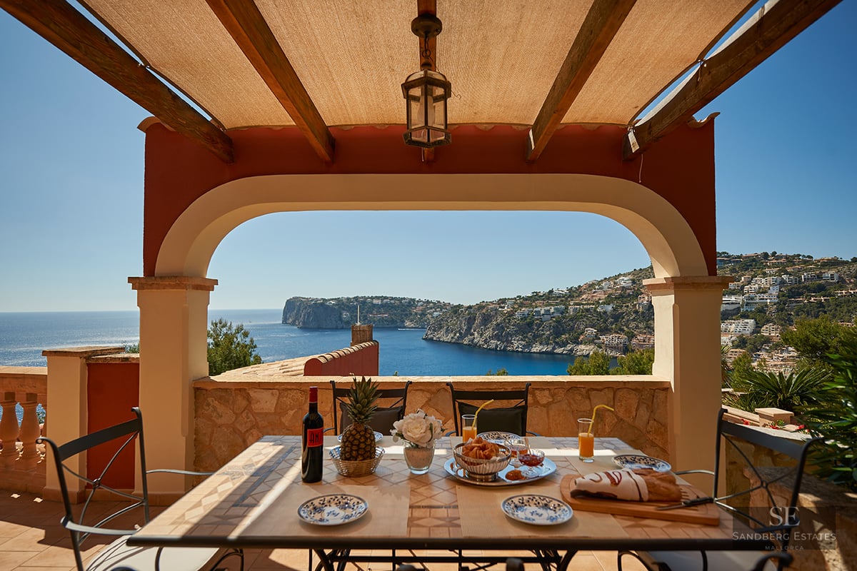 A covered Mediterranean terrace with a breakfast-set table looking out over a blue bay and coastal cliffs under a clear sky.