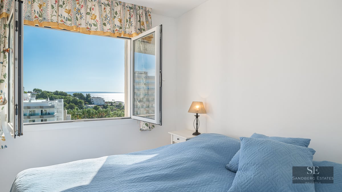 A sunlit bedroom featuring a blue quilted bed and an open window looking out over trees and the sparkling sea.