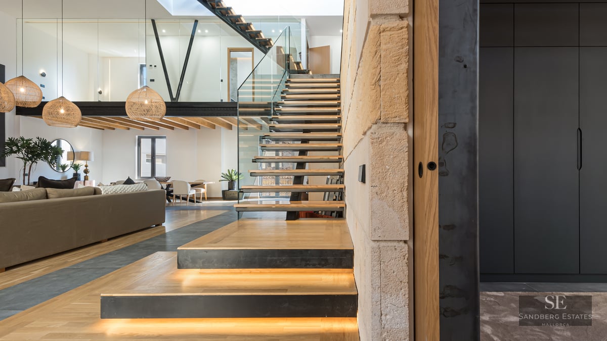Modern open-plan living room featuring a wooden floating staircase with LED lights, glass railings, and a mezzanine.