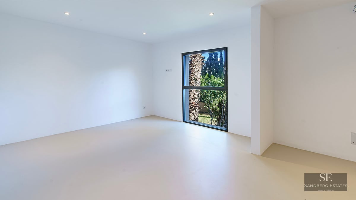Bright, empty minimalist room with white walls, light flooring, and a vertical window framing a palm tree outside.