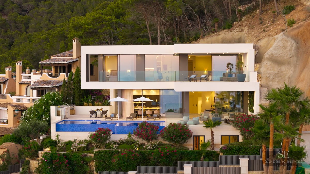 Facade of a modern white two-story villa with an infinity pool and large glass windows at dusk.