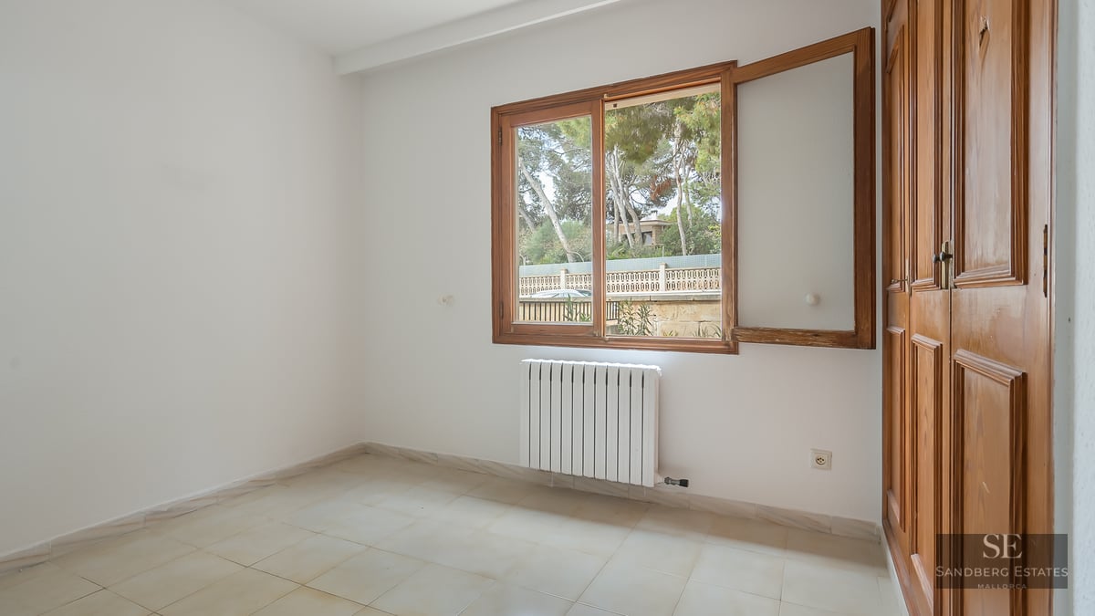 Empty bedroom with white walls, tiled floor, wooden built-in wardrobe, and window overlooking trees.