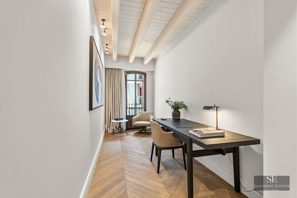 Modern home office with dark desk, chevron wood floor, white beamed ceiling, and a large window.