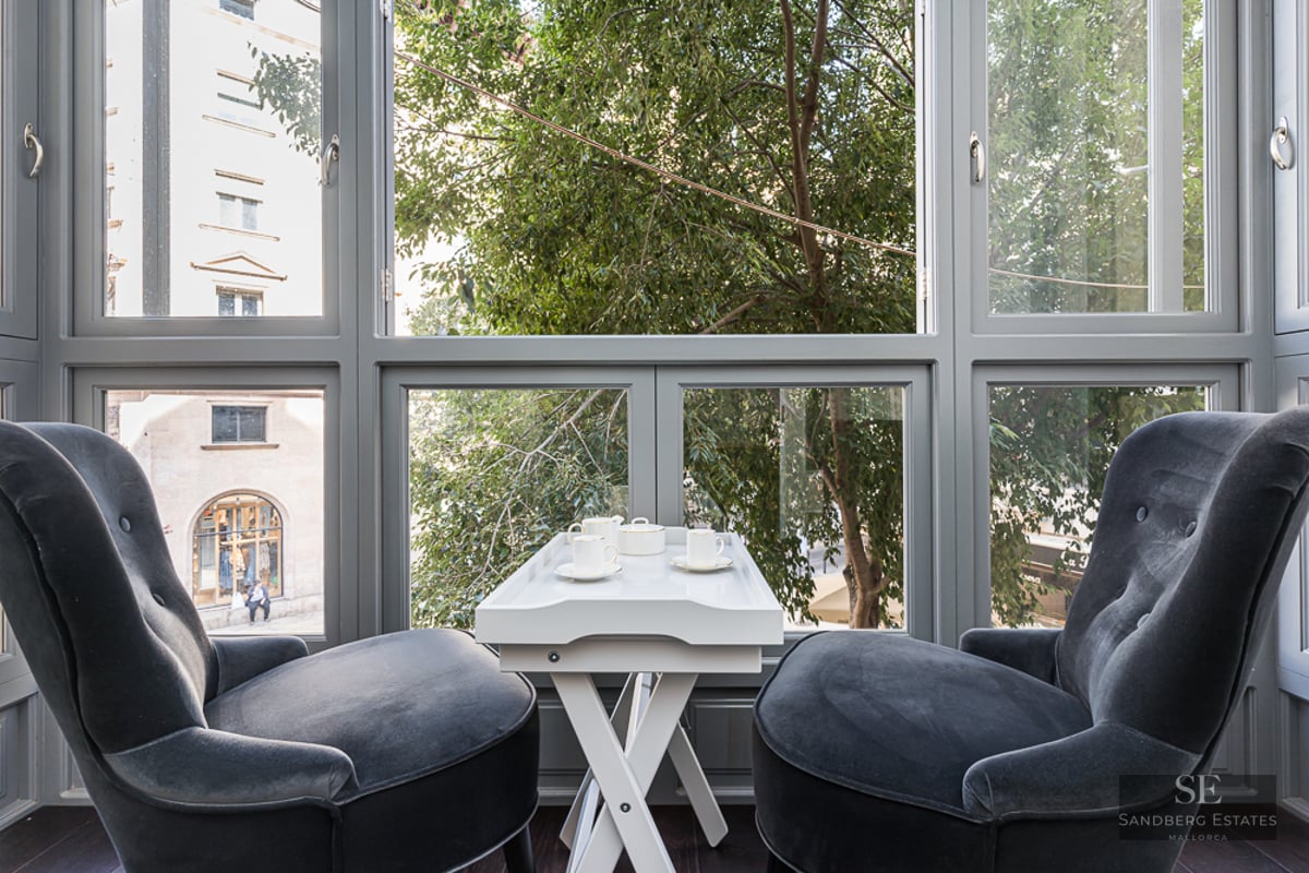 Two charcoal velvet armchairs facing a large window with a white tea table in between, overlooking city trees.