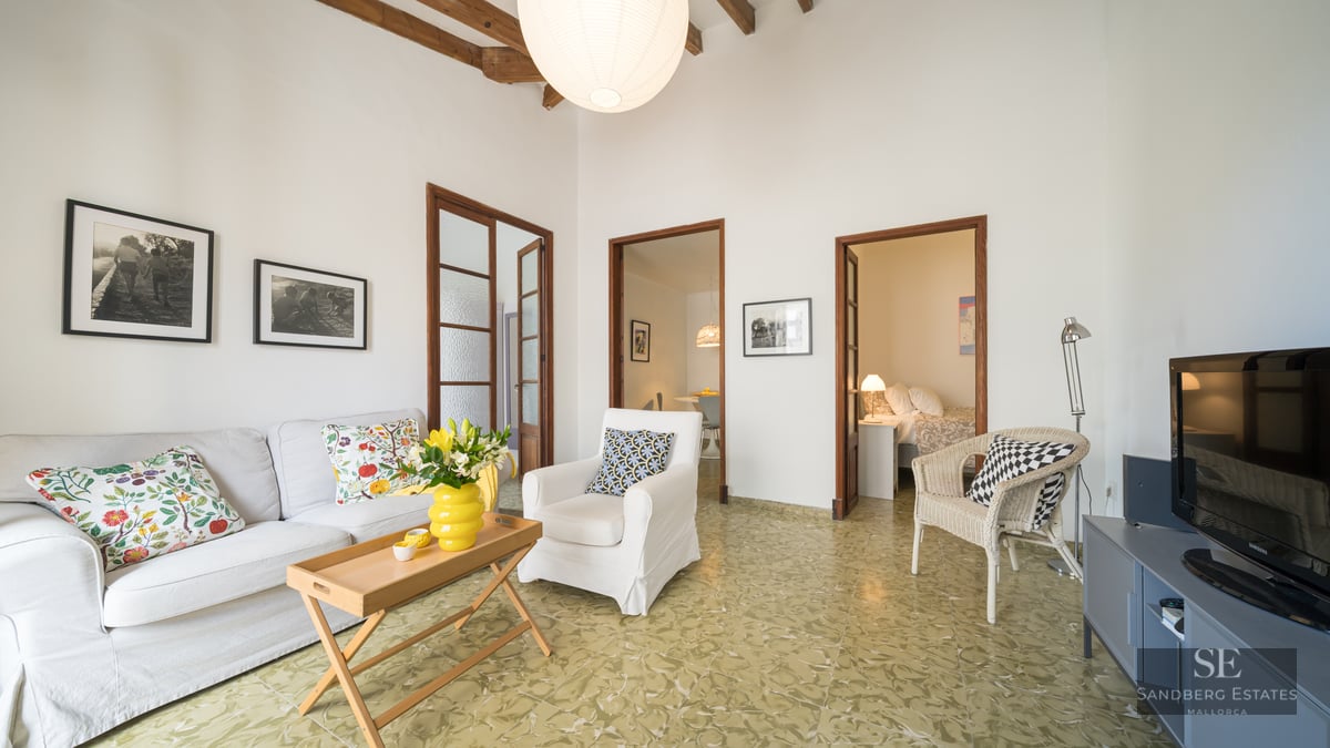 Bright living room with white sofa, rustic wooden ceiling beams, patterned green floor tiles, and doorways to other rooms.