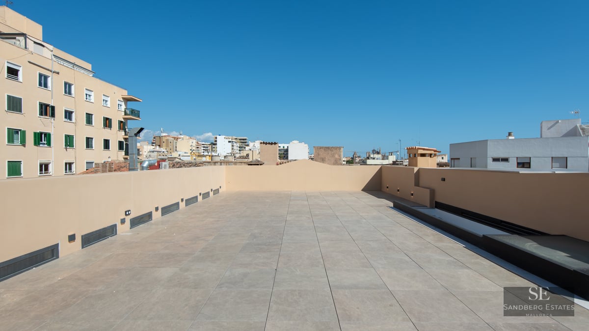 Spacious rooftop terrace with beige tiles and modern wall lighting under a clear blue sky.
