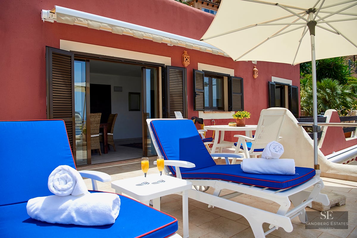 Private terrace with blue sun loungers, white towels, orange juice, and an umbrella against a terracotta building.