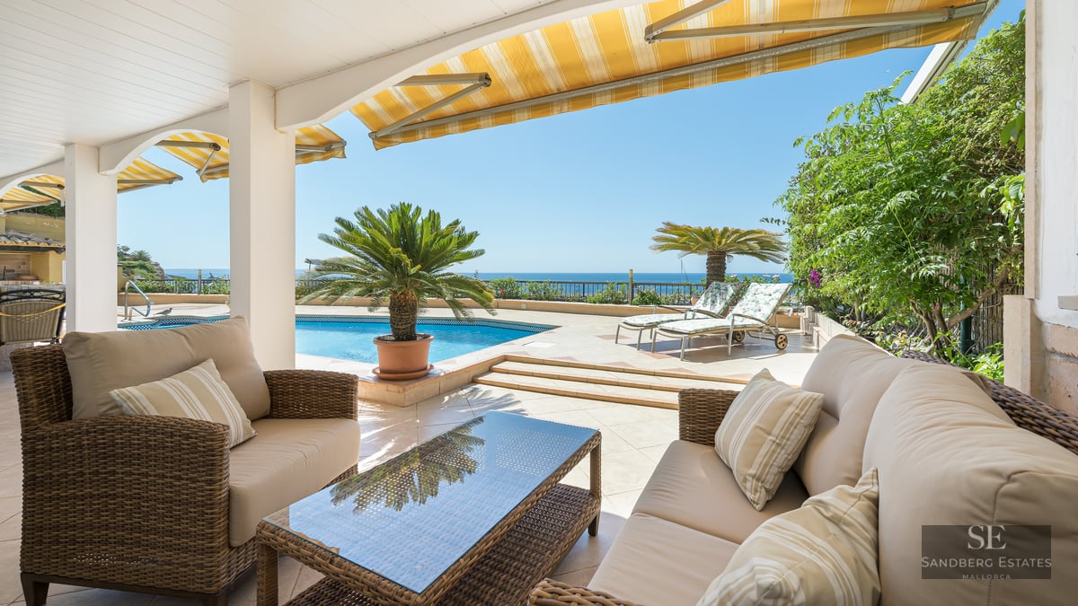 Covered terrace with wicker sofas facing a blue pool and the sea under a clear blue sky.