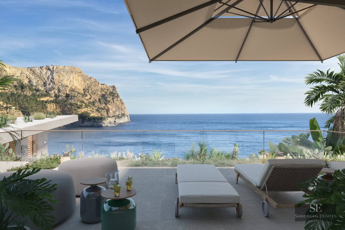 A sunny terrace with sun loungers, a parasol, and a glass railing overlooking the Mediterranean sea and a rocky cliff.