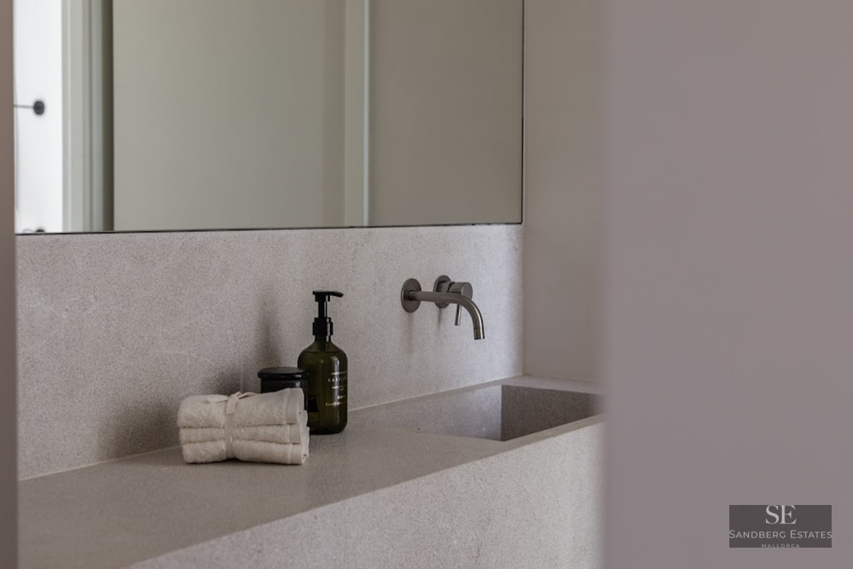 Close-up of an integrated light stone bathroom sink with a wall-mounted brushed metal faucet and luxury amenities.