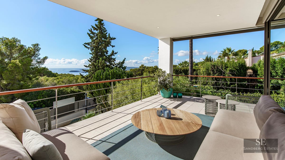 Terrasse moderne avec canapés beiges, table en bois et garde-corps métallique surplombant la mer et la verdure.