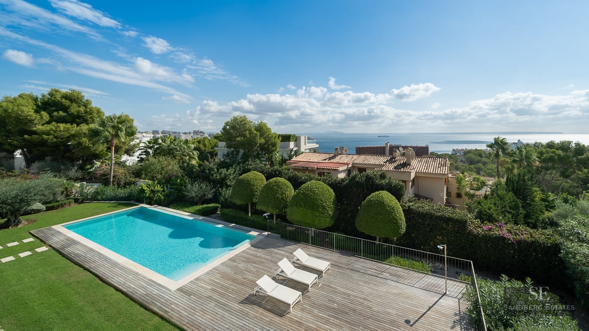 Elevated view of a rectangular turquoise swimming pool next to a wooden deck with white sun loungers and ocean views.