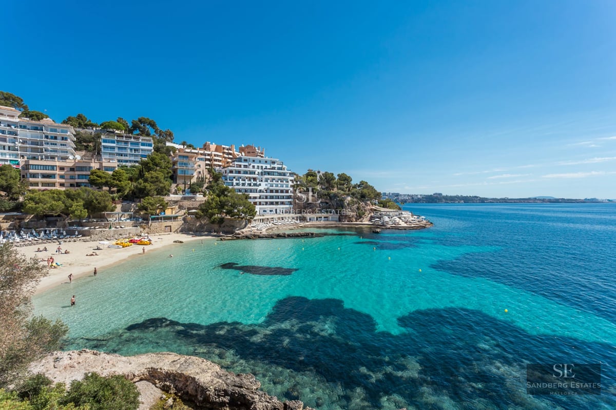 Wide panoramic view of a crystal-clear turquoise bay with a sandy beach and white buildings on a rocky coastline.