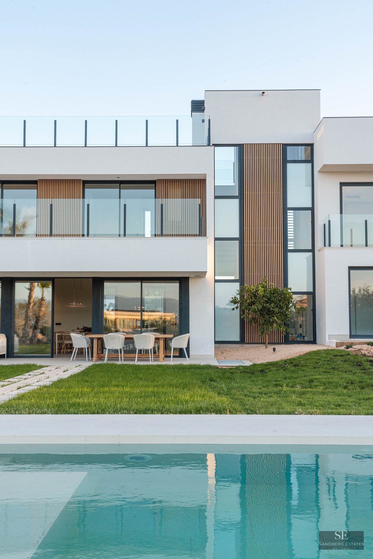 Modern white villa with wood accents, large windows, and a turquoise swimming pool in front of a green lawn.