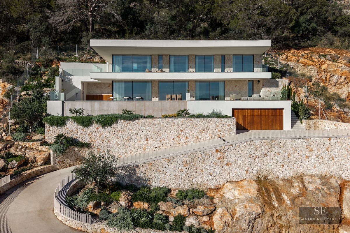 Modern white villa built into a rocky hillside with dry stone walls, large floor-to-ceiling windows, and a curved driveway.