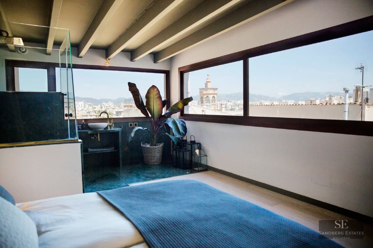 Modern bedroom with exposed wood beams, panoramic windows showing a city skyline and cathedral.