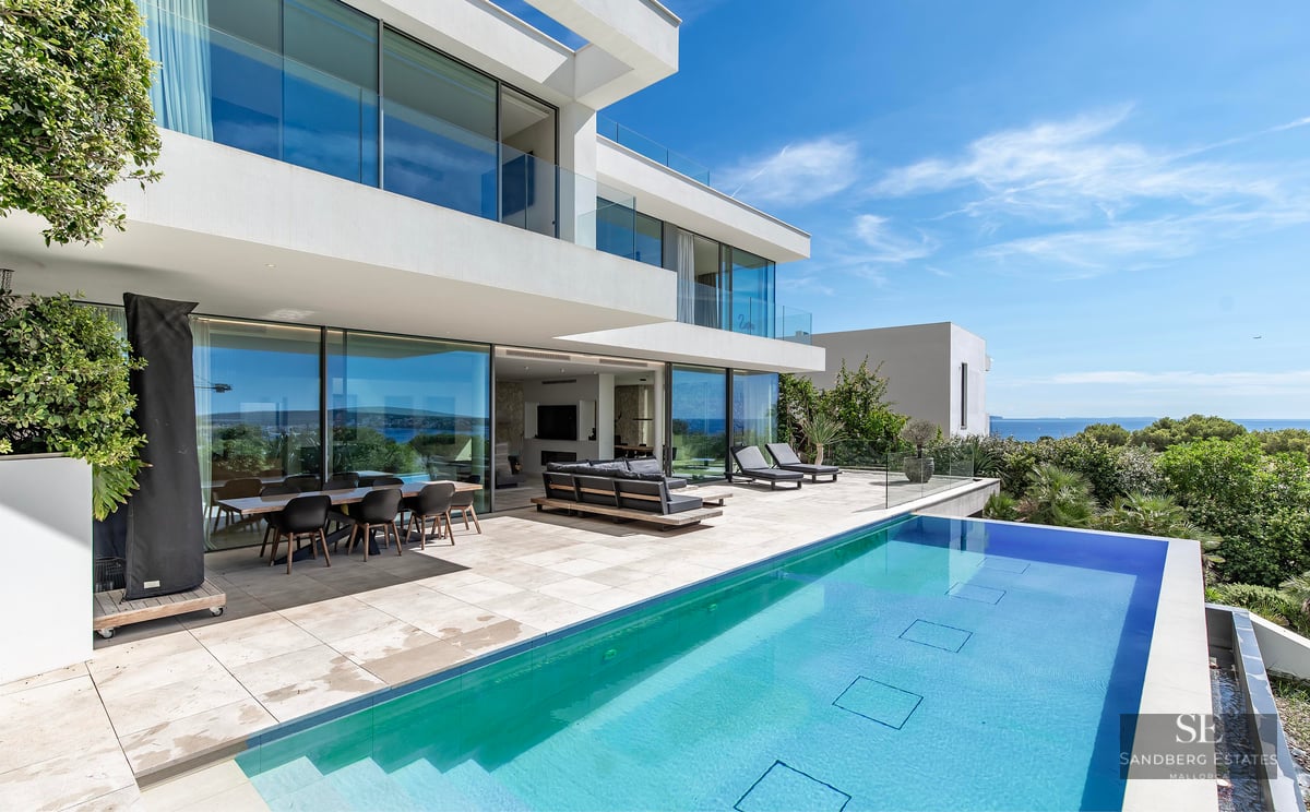 Modern white villa with an outdoor swimming pool, terrace furniture, and a panoramic view of the sea under a clear blue sky.