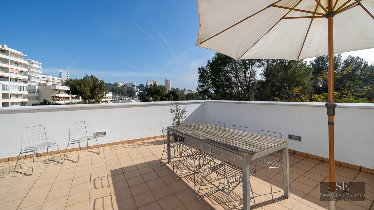 Spacious rooftop terrace with a large wooden table, wire chairs, and a white sun umbrella overlooking a city.