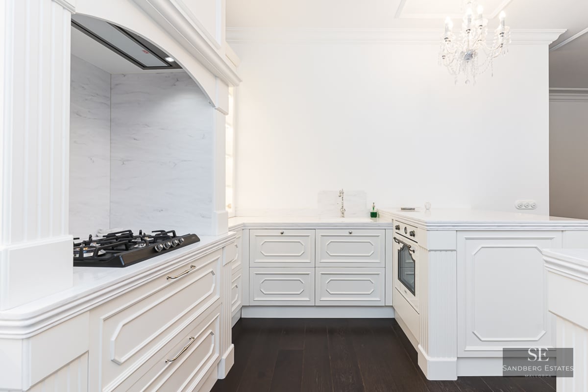 Elegant white kitchen with marble countertops, dark wood flooring, and a crystal chandelier.