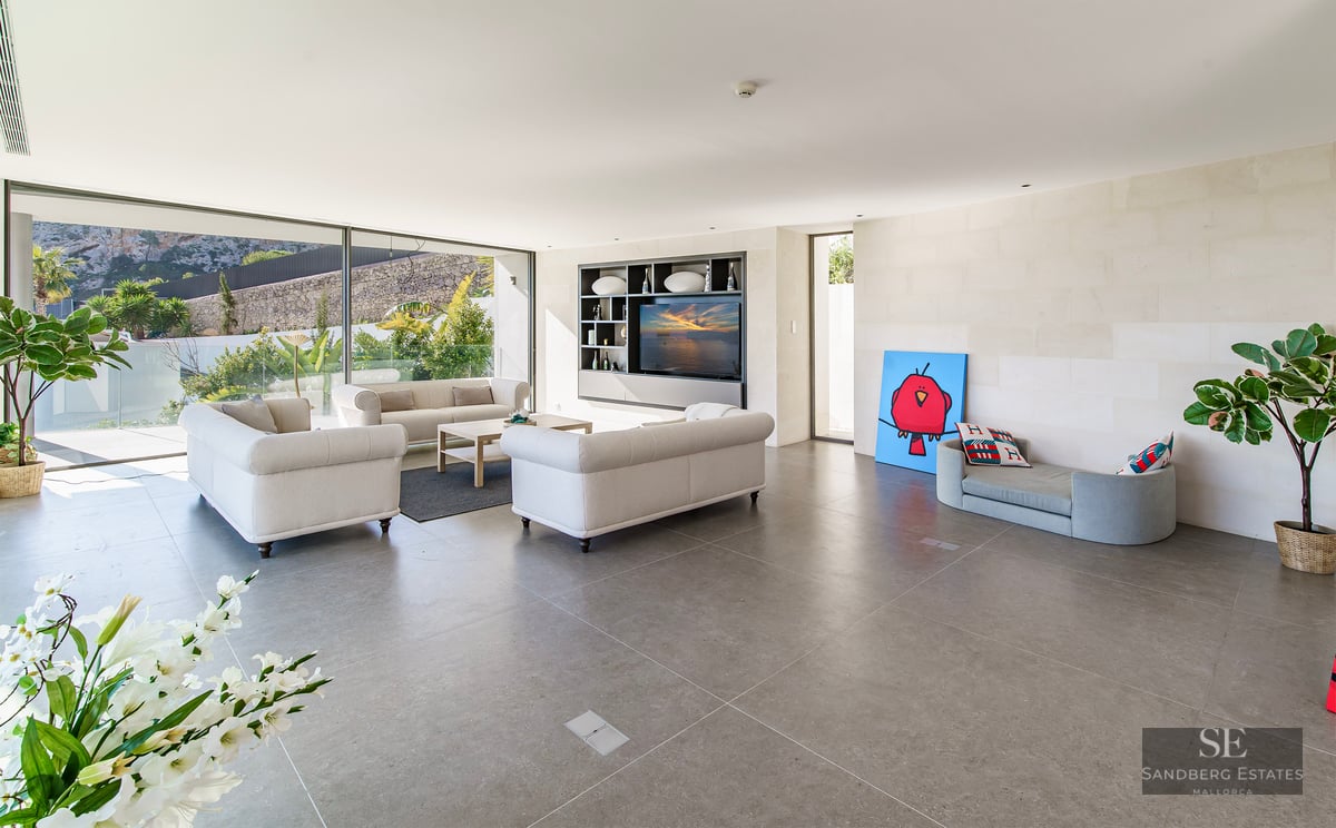 Bright modern living room with beige sofas, grey stone floor, and large glass windows overlooking a garden.
